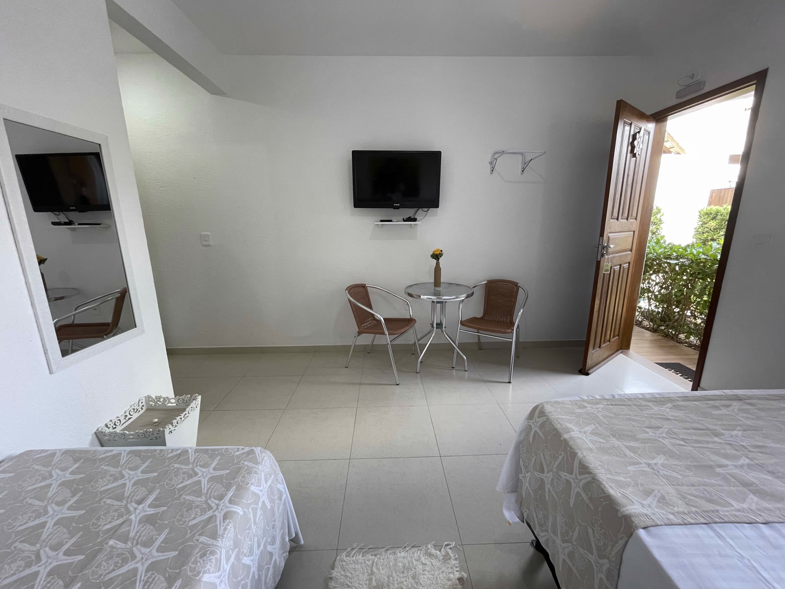 Quarto da pousada com duas camas, mesinha com duas cadeiras, TV na parede e a porta aberta para o jardim